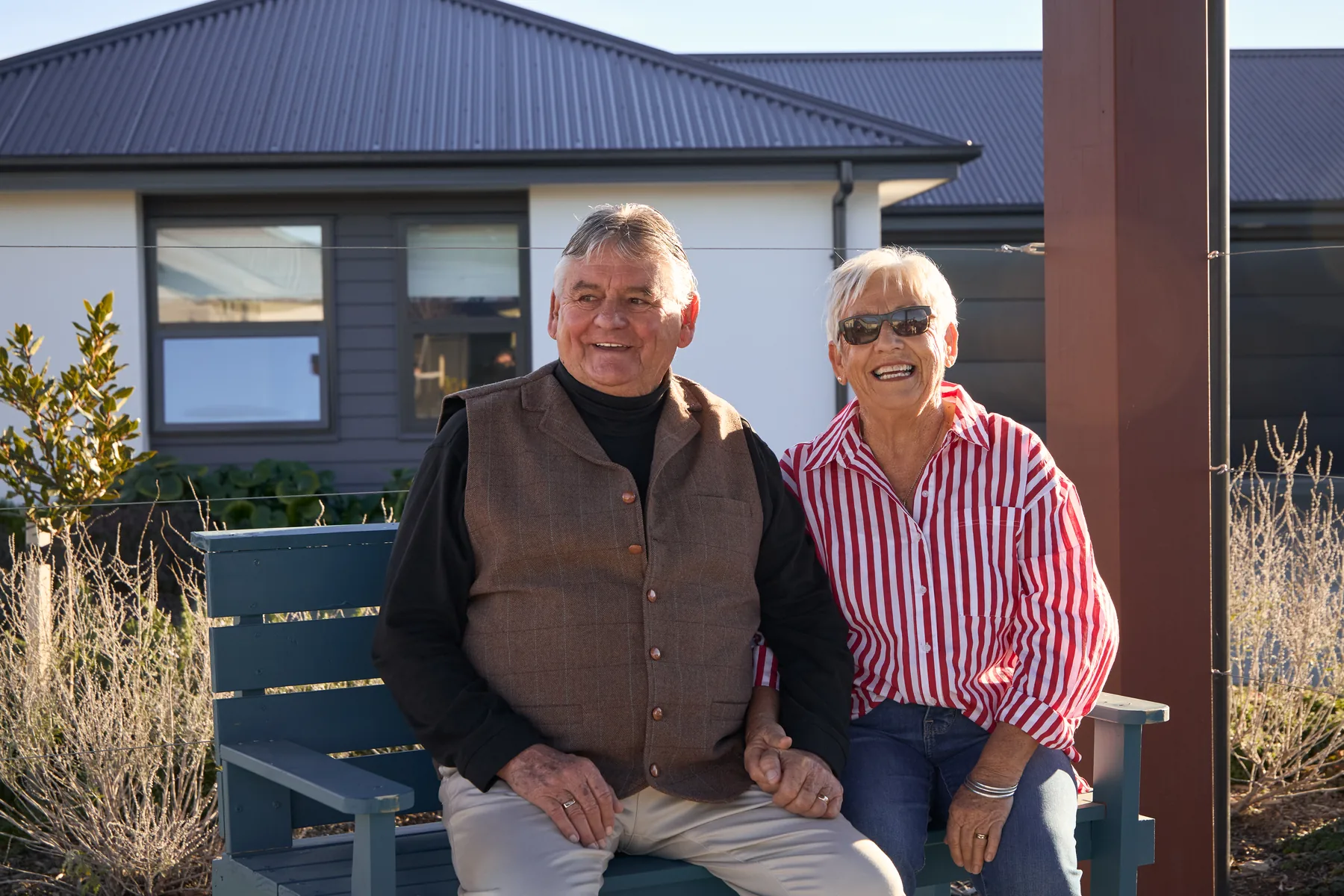 A man and a woman sit side by side on a bench outside a house, both smiling.